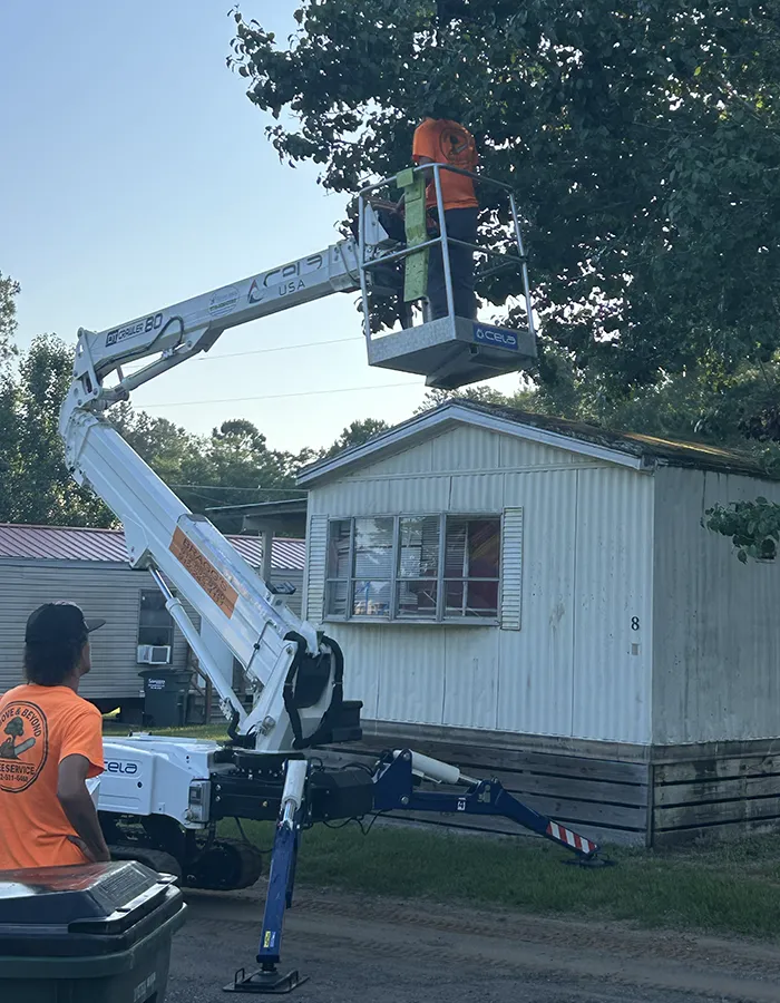 Worker in orange shirt stands in elevated bucket of a boom lift trimming branches beside a white mobile-home.